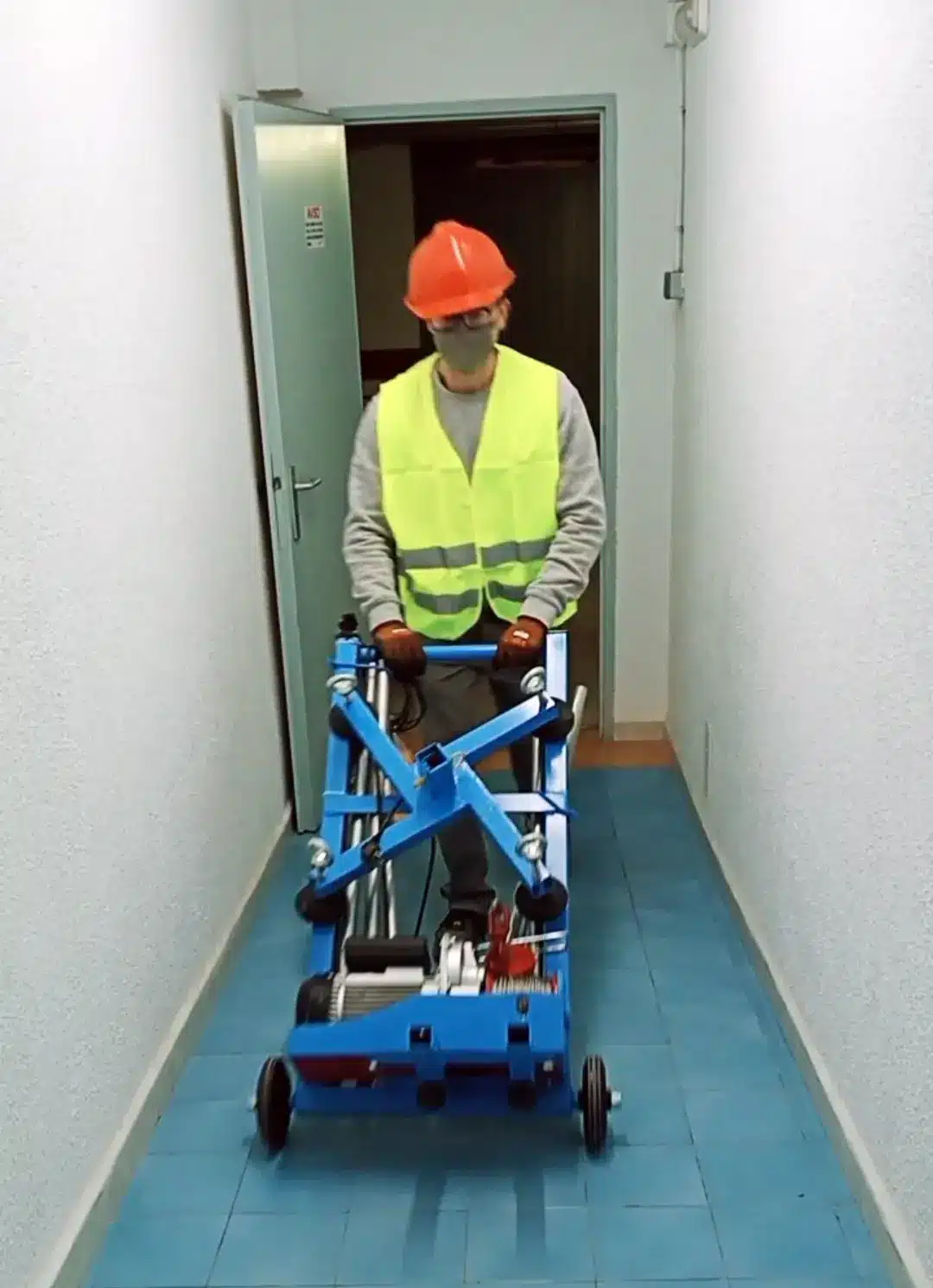 Työntekijä kuljettaa taittuvaa Elevall 400 -kerrosnosturia kapeassa käytävässä; laite mahtuu oviaukosta ja liukuu pyörillä.