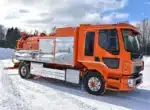 Oranssi Volvo-alustainen imupaineauto lumisella tiellä, säiliön kyljessä ruostumattomasta teräksestä valmistetut kaapit ja perässä imuletkuvarusteet.